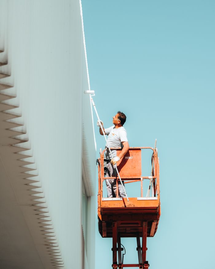 gallery-1 A man painting a building exterior from a cherry picker against a clear blue sky.