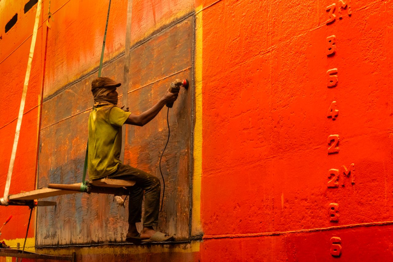 hero-img-01 A worker carefully sands the vibrant orange hull of a ship, engaged in maritime maintenance.