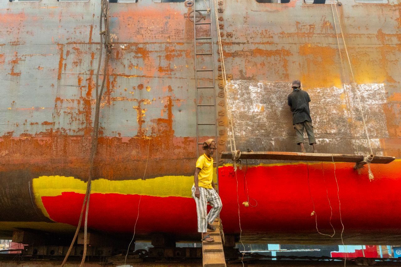 services-02 Two workers refurbishing a rusty ship hull with bright paint, showcasing industrial shipyard labor.