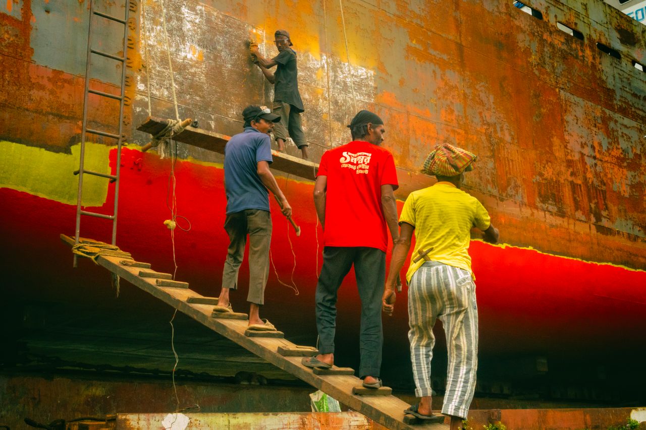 hero-img-02 Four workers refurbishing the hull of a rusted ship in a dockyard, highlighting teamwork and labor.