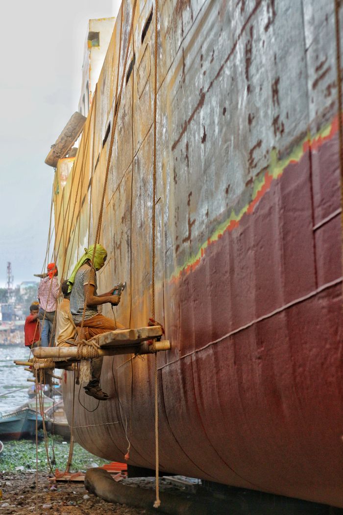 gallery-2 Industrial laborers painting the hull of a ship at a shipyard, showcasing hard work and craftsmanship.