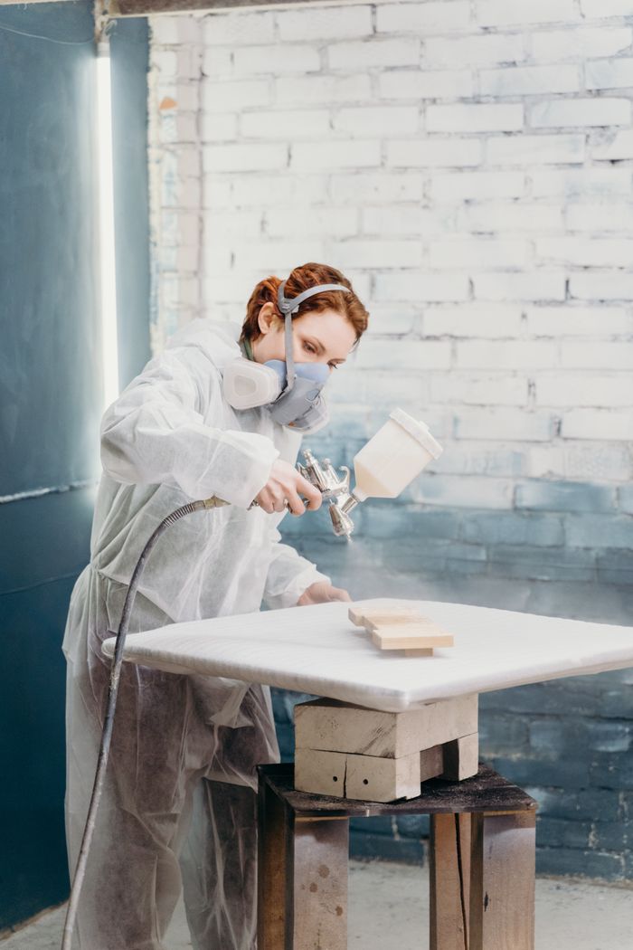 gallery-3 A woman in protective gear spray paints a table in a modern workshop environment.